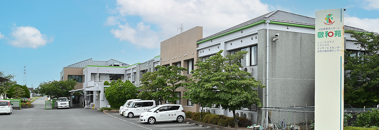 介護施設の建物外観。複数棟の平屋から2階建て建物が見え、駐車場と緑化された敷地を備えている。