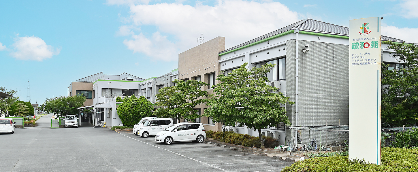 介護施設の建物外観。緑のラインが特徴的な白色建物と駐車場