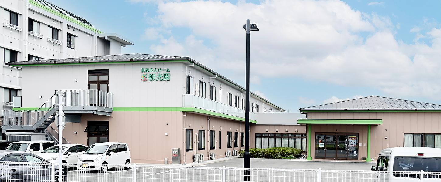 介護施設の建物外観。白とピンク色の壁、緑色の屋根が特徴的な施設。駐車場も見える。